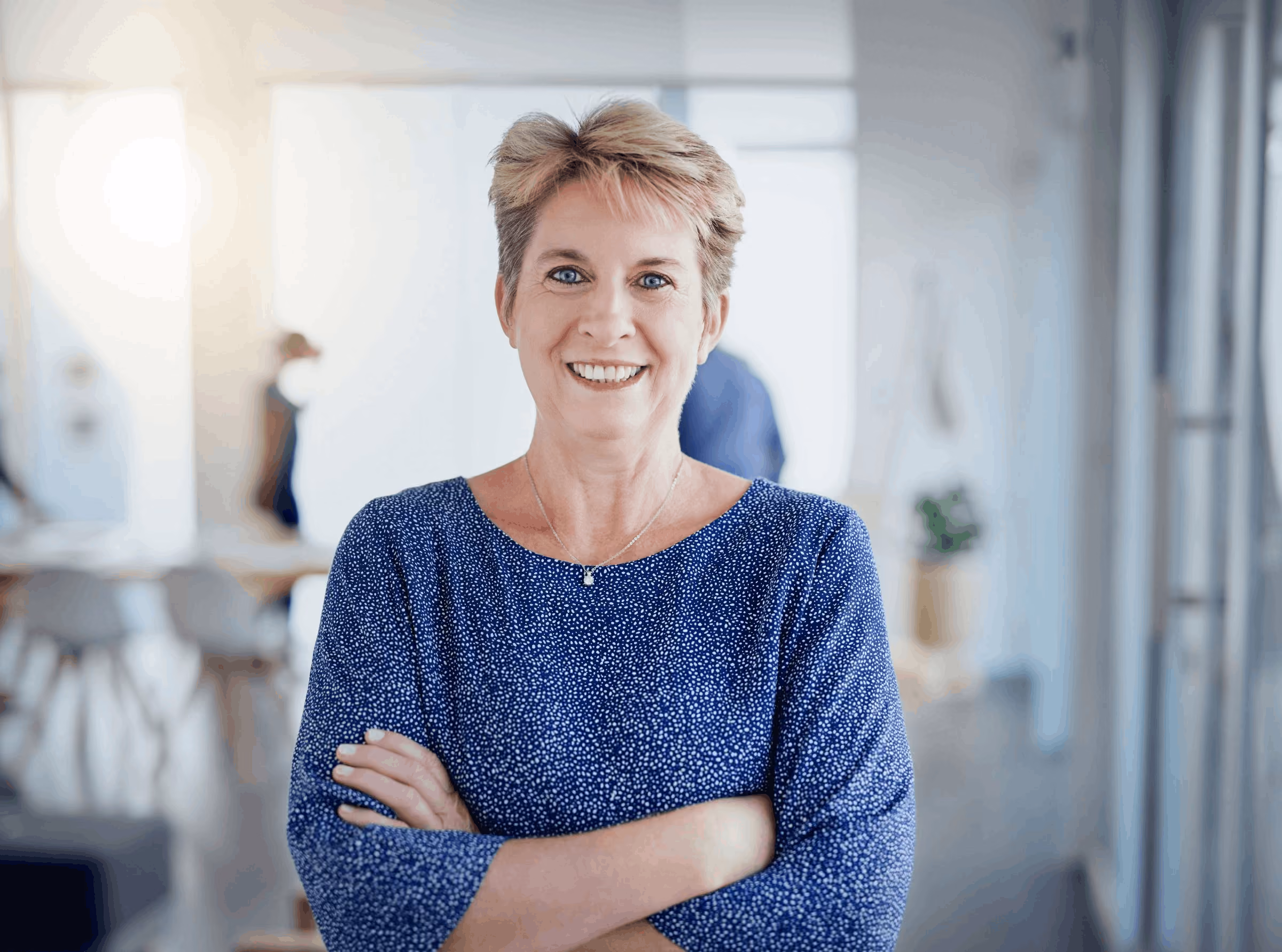 Smiling middle-aged woman with short blonde hair wearing a blue patterned top with arms crossed indoors.