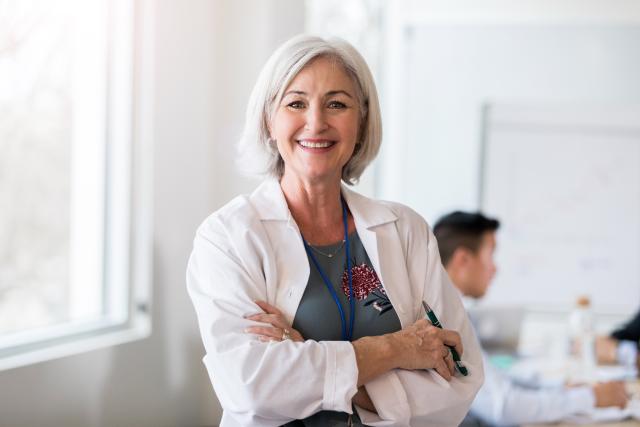 Smiling middle-aged woman in a white lab coat standing with arms crossed in a bright room.