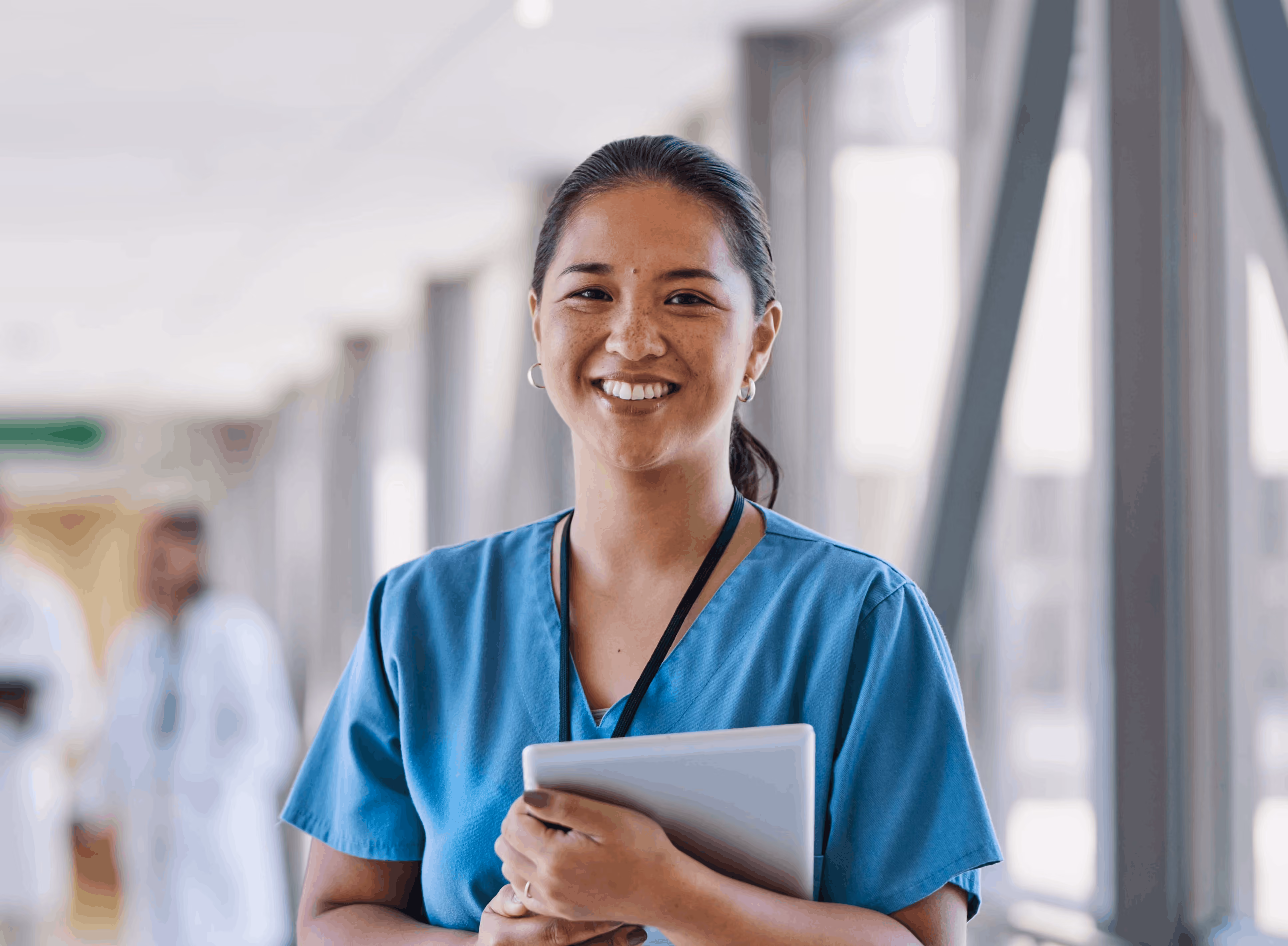 Smiling female nurse in blue scrubs holding a tablet in a hospital corridor.