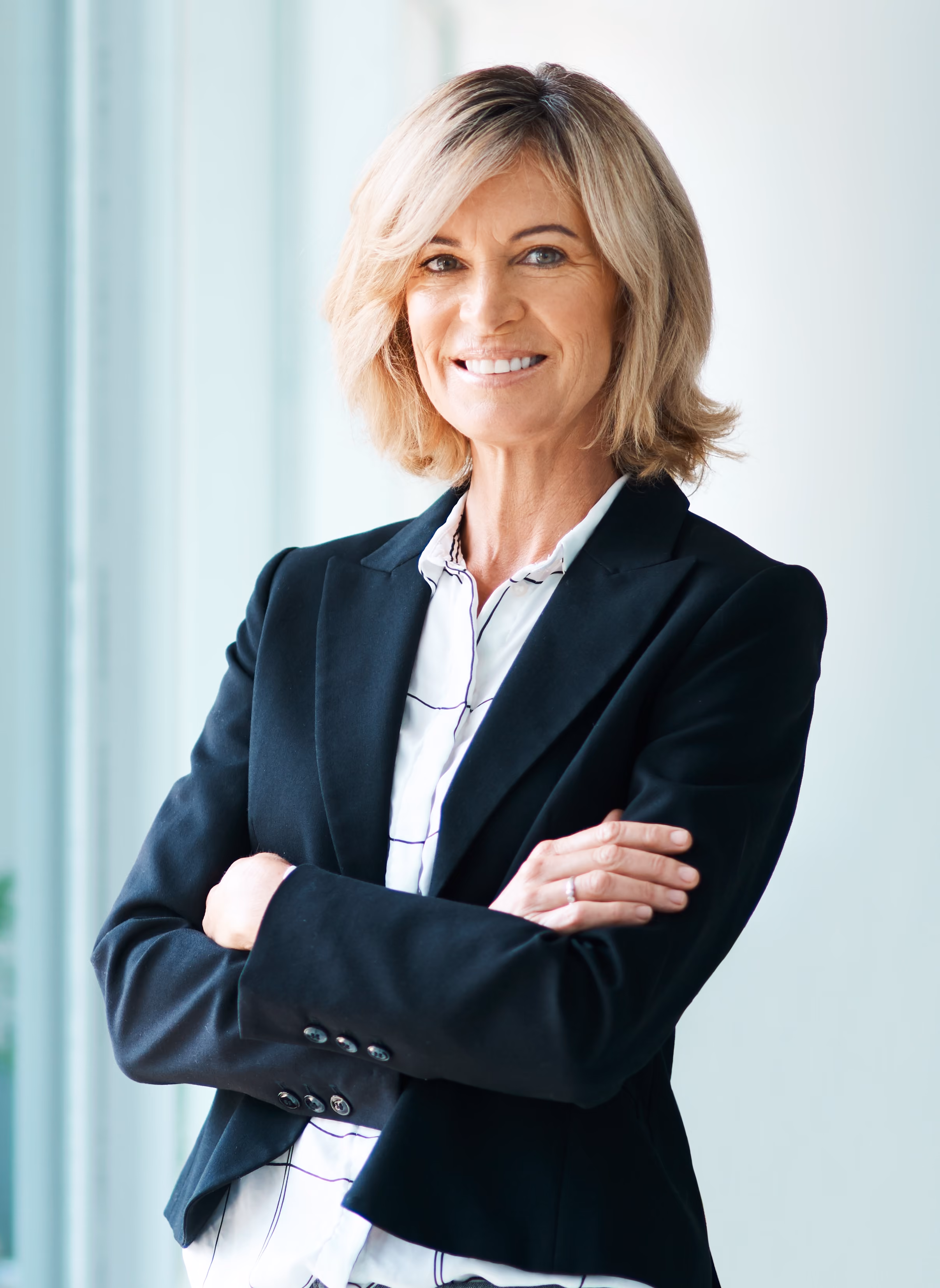 Confident middle-aged woman with blonde hair, wearing a black blazer and white shirt, arms crossed.