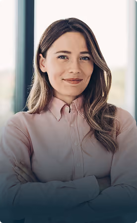 Confident woman with long brown hair wearing a light pink button-up shirt standing with arms crossed, smiling slightly against a blurred window background.