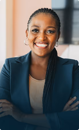 Smiling Black woman with braided hair wearing hoop earrings and a dark blazer, standing with arms crossed indoors in front of blurred window.
