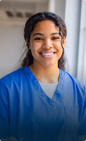Smiling young nurse or healthcare professional with dark curly hair wearing blue scrubs standing indoors near a window.