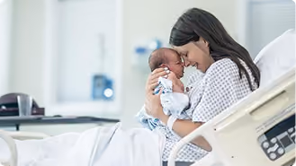 A woman in a hospital gown sitting on a hospital bed, holding and gently touching foreheads with a newborn baby wrapped in a blanket.