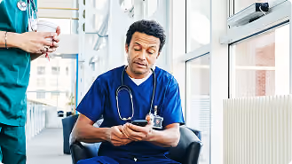 Male healthcare professional in blue scrubs sitting and looking at his phone in a bright hospital corridor, with another person in green scrubs standing nearby holding a cup.