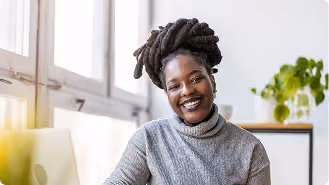 Smiling woman with dreadlocks wearing a gray turtleneck sitting indoors near a window with a potted plant in the background.