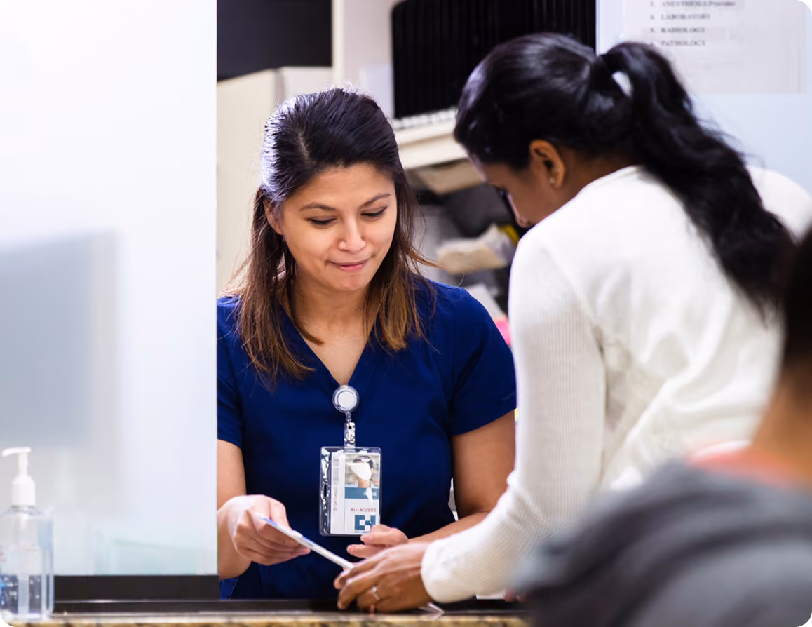 Female healthcare worker in a navy blue scrub top with an ID badge handing a document to a woman with dark hair tied back, standing at a counter in a medical or office setting.