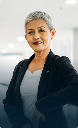 Confident older woman with short gray hair wearing a black blazer and white top, standing in a modern office setting.