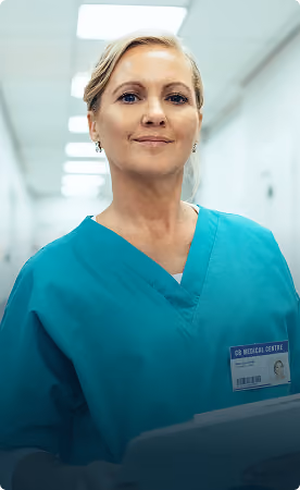 Confident female medical professional in turquoise scrubs holding a tablet, standing in a hospital corridor with bright overhead lights.