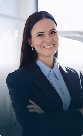 Portrait of a smiling woman with long dark hair wearing a dark blazer and light blue blouse, standing with crossed arms in a bright office setting.