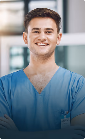Smiling young male healthcare professional wearing blue scrubs with arms crossed, standing in a bright hospital setting.