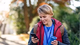 Smiling woman outdoors looking at her phone, wearing a maroon and black jacket with a backpack, holding a blue cup.