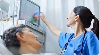 A female healthcare professional in blue scrubs adjusting a monitor mounted on a wall next to a patient lying in a hospital bed.