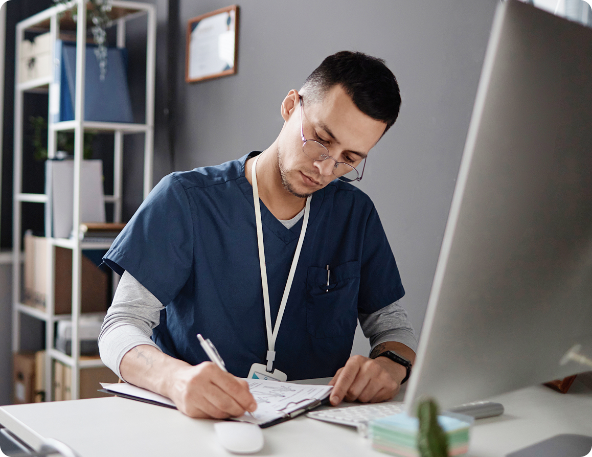 Medical professional wearing navy scrubs and glasses writing on a clipboard at a desk with a computer monitor and keyboard.