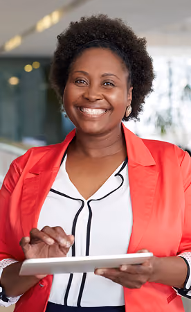 Smiling woman with short curly hair wearing a red blazer and white blouse, holding and using a tablet in an indoor office setting.