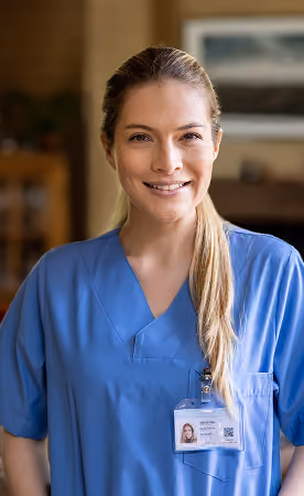 Smiling female healthcare worker wearing blue scrubs and an ID badge, standing indoors with blurred background.