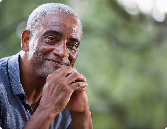 Smiling mature man with gray hair, resting his chin on his hands, with a blurred green outdoor background.