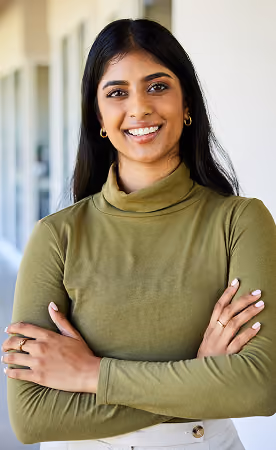 Smiling young woman with long dark hair and gold hoop earrings, wearing an olive green turtleneck and crossing her arms.
