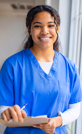 Smiling female healthcare professional wearing blue scrubs and holding a clipboard and pen near a window with natural light.