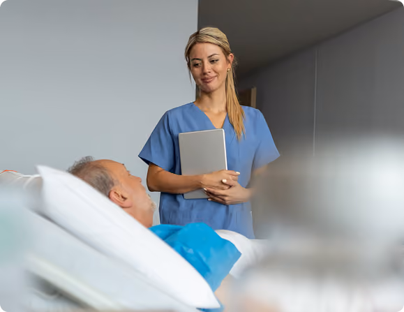 A nurse in blue scrubs holding a tablet stands beside a hospital bed, smiling at an elderly male patient lying in bed with white pillows.