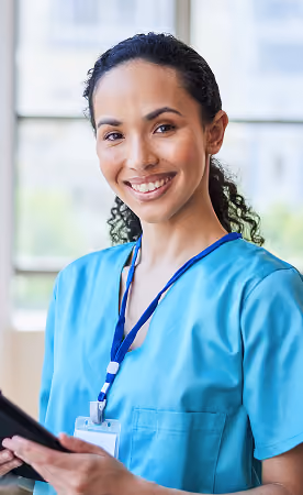 Smiling healthcare professional in blue scrubs holding a tablet with a lanyard and ID badge around her neck, standing indoors near a window.