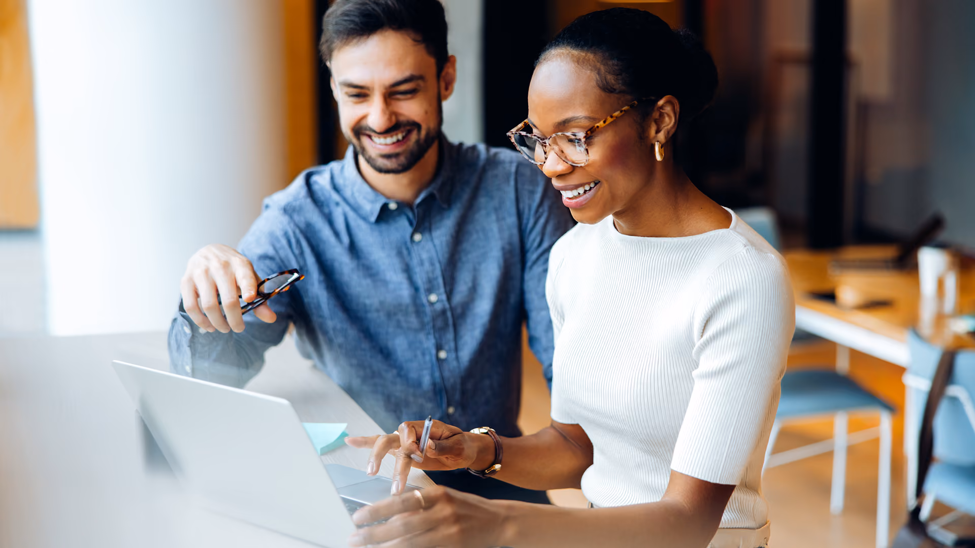 Smiling man and woman sitting at a desk working together on a laptop in a bright office space.