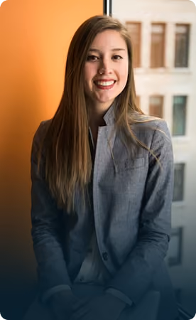 Smiling young woman with long brown hair wearing a gray blazer sitting indoors near a window with an orange wall in the background.