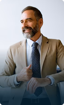 Smiling middle-aged man with a beard wearing a beige suit jacket, white shirt, and dark blue polka dot tie, looking to the side against a plain light background.