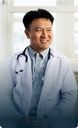 Smiling male doctor wearing a white coat and stethoscope around his neck sitting in front of a window.