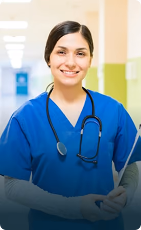 Smiling female healthcare professional wearing blue scrubs and a stethoscope around her neck, standing in a hospital corridor.