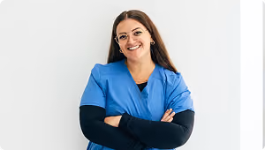 Smiling healthcare worker wearing blue scrubs and glasses, standing with arms crossed against a white background.