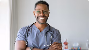 Smiling male healthcare professional wearing gray scrubs and glasses with a stethoscope around his neck, standing with arms crossed.