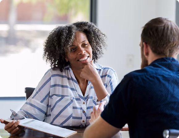 A woman with curly hair wearing a striped shirt smiles while holding a clipboard during a conversation with a man seated across from her.