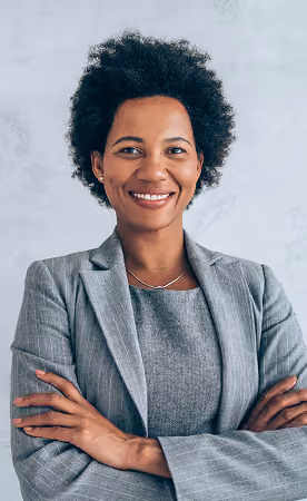Smiling professional Black woman with natural curly hair wearing a gray business suit and arms crossed against a light background.