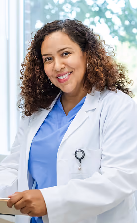 Smiling female healthcare professional with curly hair wearing blue scrubs and a white lab coat, standing indoors near a window.