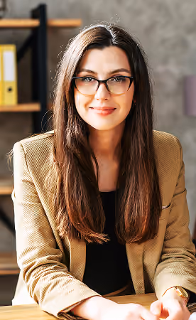 Young woman with long brown hair and glasses wearing a tan blazer and black top sitting at a desk, smiling softly toward the camera in a modern office setting.