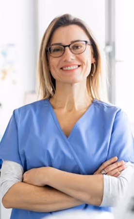 Smiling woman with glasses wearing blue medical scrubs and a light long-sleeve shirt underneath, standing with arms crossed in a bright, blurred indoor setting.