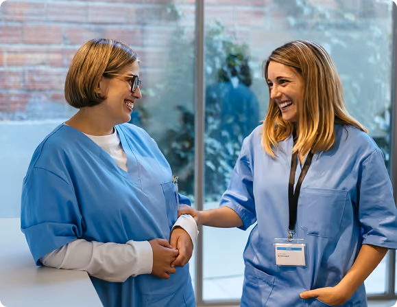 Two female healthcare workers in blue scrubs smiling and talking indoors by a large window, one resting her hand on the other's arm.