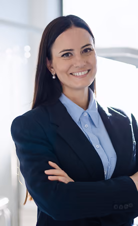 Smiling woman with long dark hair wearing a dark blazer and light blue shirt, standing with arms crossed in a bright office setting.