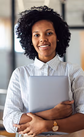 Smiling woman with curly hair wearing a striped white blouse, holding a silver laptop in a modern office setting.