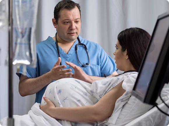 A male doctor in blue scrubs with a stethoscope talks to a pregnant woman lying in a hospital bed.