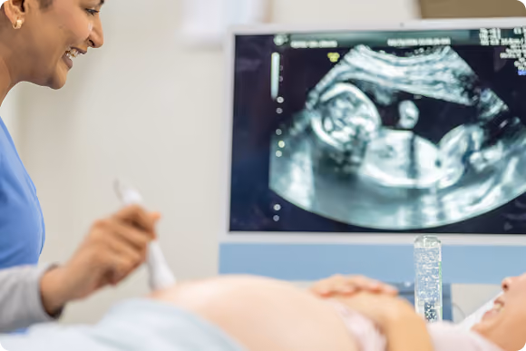 Pregnant woman lying on a medical exam table smiling while a healthcare professional performs an ultrasound, with the ultrasound image of the fetus shown on a monitor nearby.