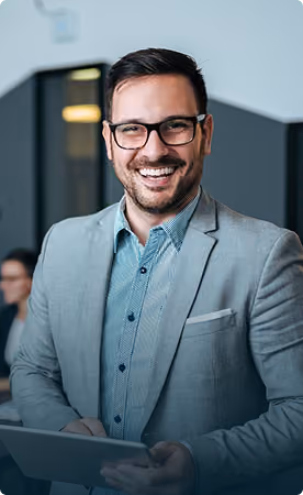 Smiling man wearing glasses, a light blue button-up shirt, and a grey blazer, holding a tablet in a modern office setting.