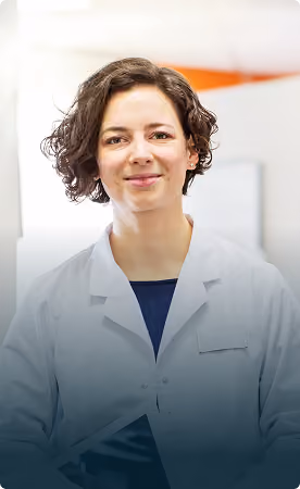 Smiling female physician with curly brown hair wearing a white lab coat and dark blue shirt standing in a clinical or hospital environment.