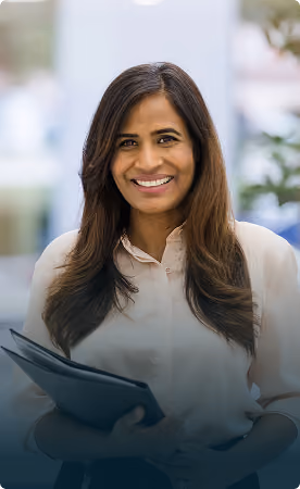 Smiling woman with long dark hair wearing a light-colored shirt, holding a folder, standing indoors.