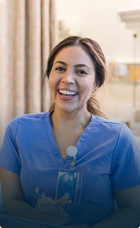 Smiling female nurse wearing blue scrubs and ID badge, holding a pen and clipboard in a clinical setting.