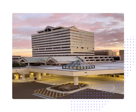 Large multi-story office building at sunset with a lower attached structure featuring a glass pyramid skylight and surrounding parking area.