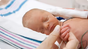 Close-up of a premature newborn baby resting with eyes closed, wrapped in a striped hospital blanket, with a hand gently holding the baby's tiny hand, and medical tubes attached.