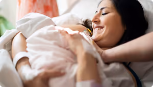 Smiling woman lying in bed gently holding and looking at a baby wrapped in a soft blanket.