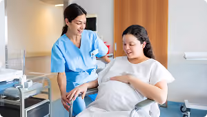 Pregnant woman sitting in a hospital chair holding her belly, smiling as a nurse in blue scrubs stands beside her with a comforting hand on her shoulder.